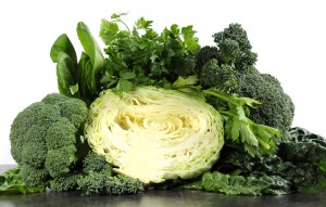 Healthy diet health foods with leafy green vegetables including cabbage, broccoli, broccolini, parsley, celery, silverbeet, and spinach on black slate kitchen top and white wall.
