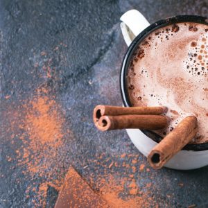 Vintage mug of hot chocolate with cinnamon sticks over dark background. Top view. Square image with selective focus
